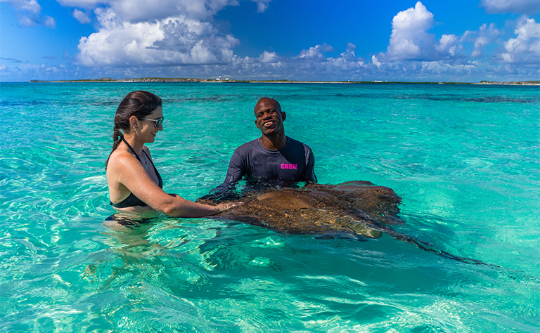 A woman and a guide standing in shallow turquoise water interacting with a stingray under a bright, partly cloudy sky.